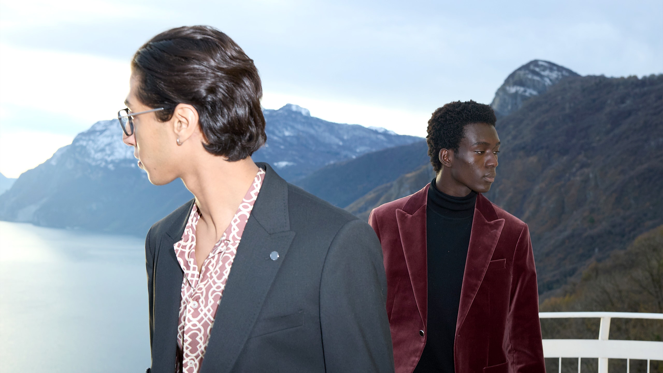 Two men in stylish blazers stand near a railing with a mountainous lake landscape in the background.