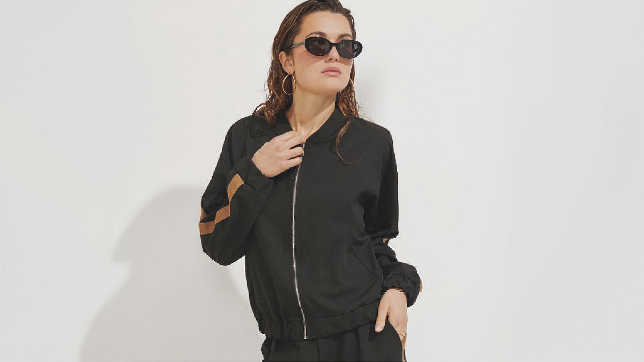 Woman wearing black sunglasses and a black zip-up jacket with brown stripes on the sleeves.